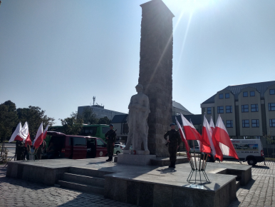Na zdjęciu widzimy dwóch żołnierzy z karabinami, dwa stanowiska z flagami Rzeczypospolitej Polskiej oraz pomnik Żołnierza Polskiego; w tel błękitne niebo i promienie słońca spadające na pomnik